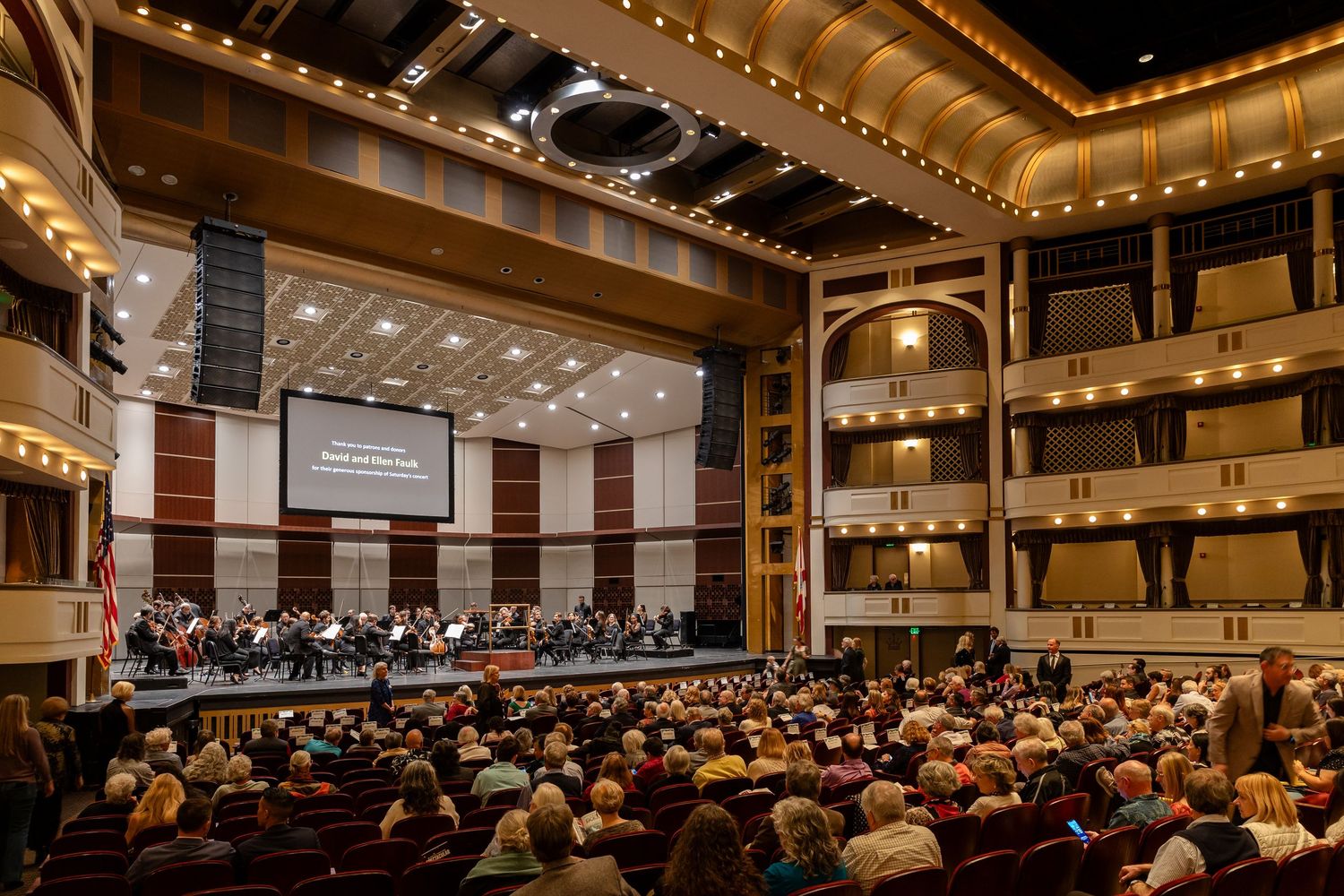 Orchestra performing on stage in an elegant concert hall filled with audience members seated in red chairs.