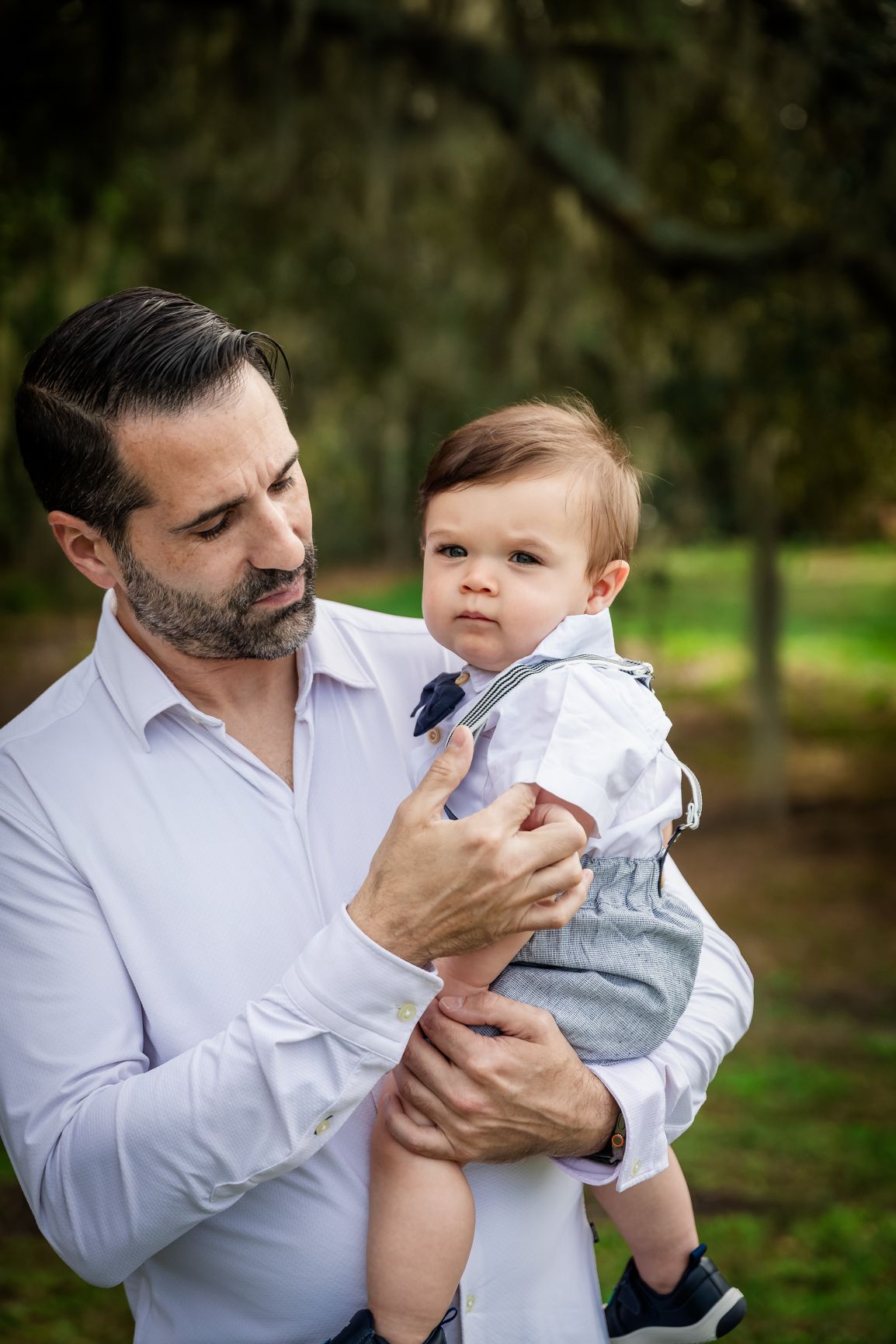 Father in white shirt tenderly holding infant son outdoors under moss-covered trees.
