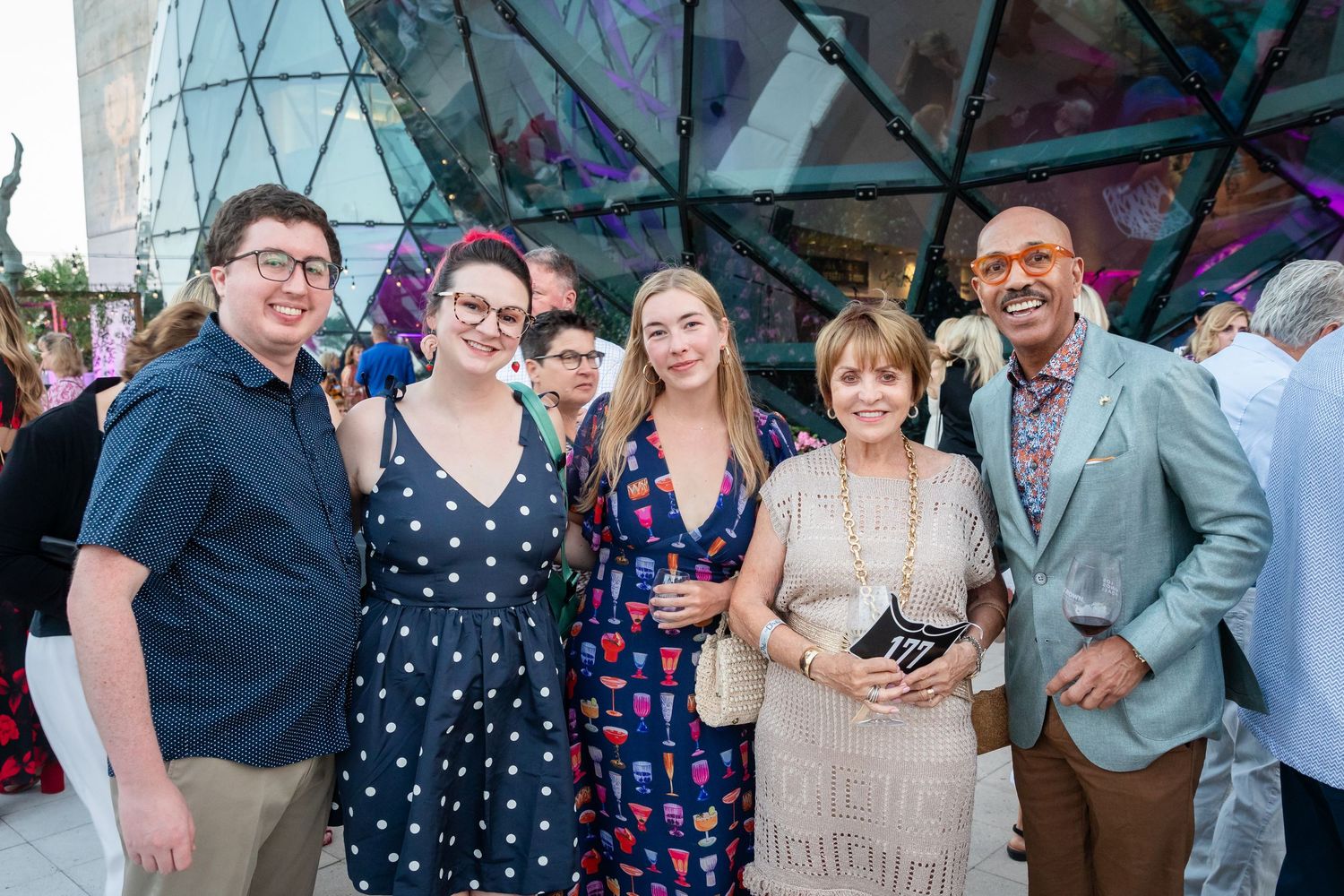 Group of six people smiling together at an elegant outdoor evening event with colorful decorative backdrop.