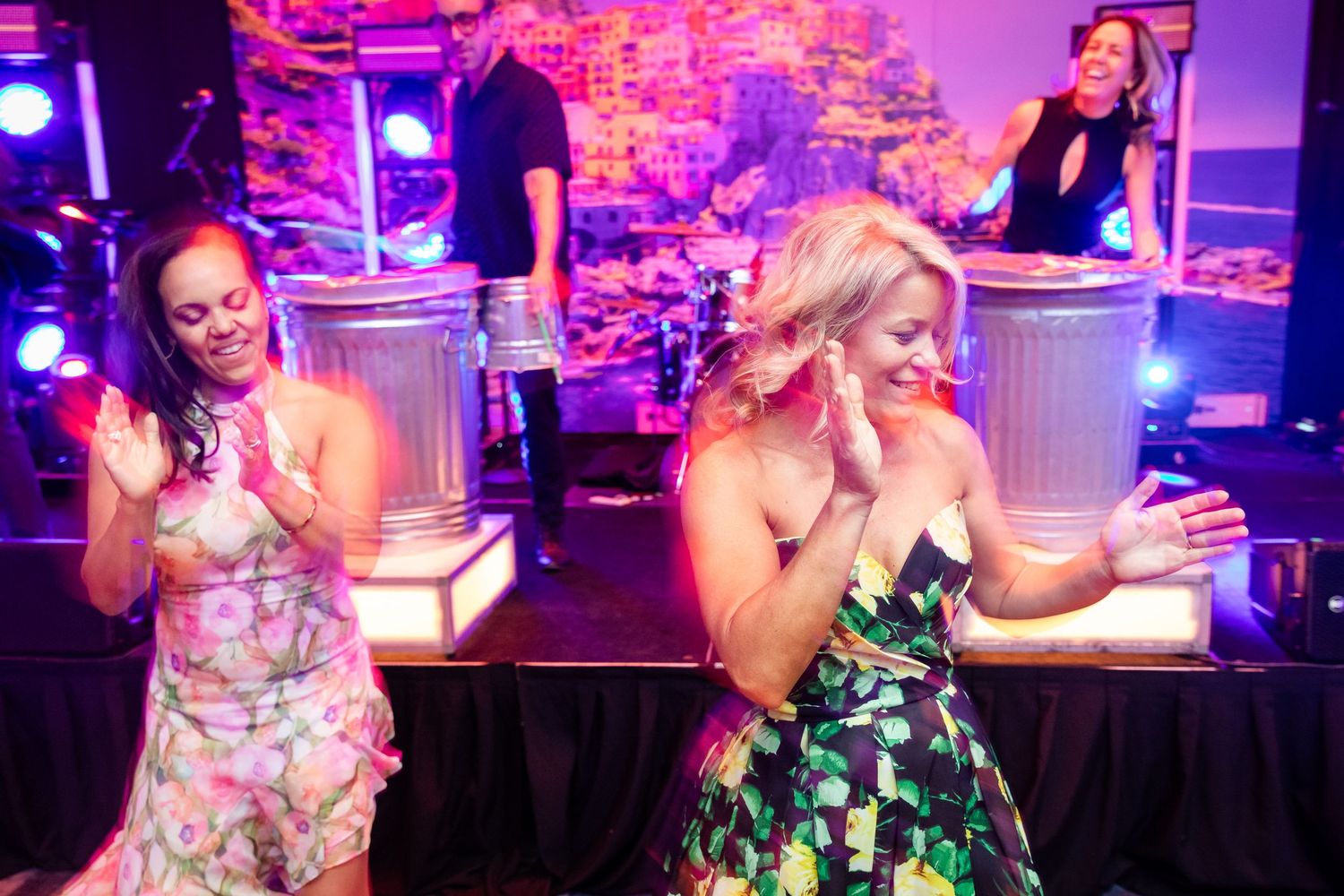 Two women dancing energetically at a lively event with a DJ performing on illuminated trash cans on stage.