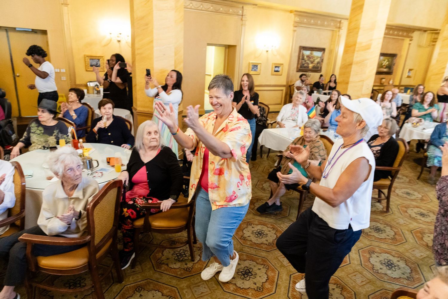 Crowd of seniors clapping and dancing at a lively indoor celebration event in a grand ballroom.