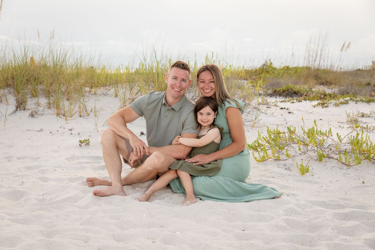 Family of three sitting on white sand beach, parents smiling with child, wearing coordinated green outfits near sand dunes.