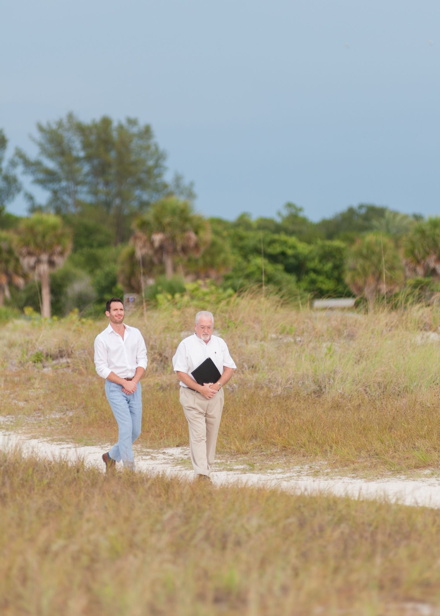 Two men walking along a sandy path through coastal grassland with palm trees, one carrying a black folder.