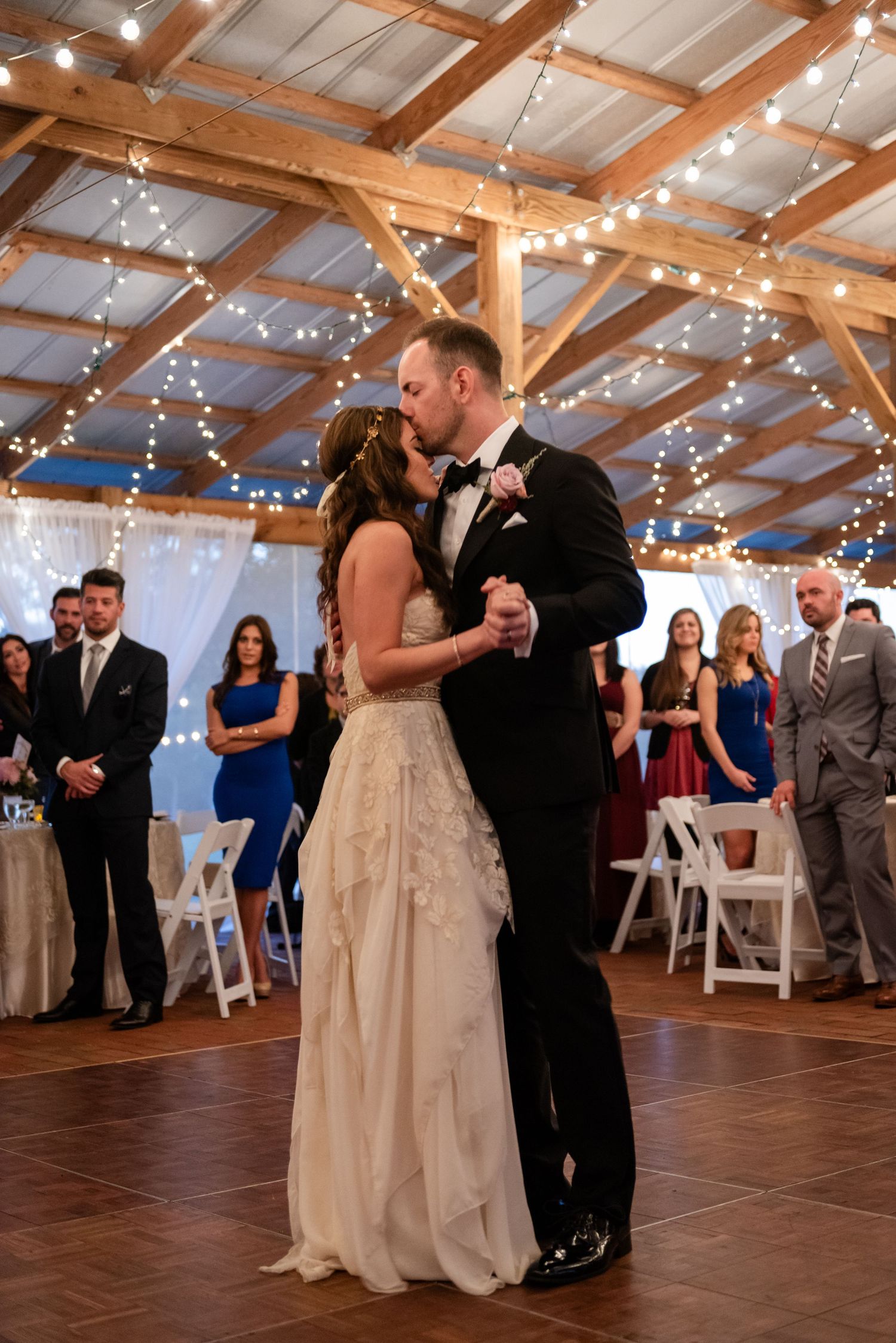 Bride and groom share first dance in rustic barn venue with string lights and wedding guests.