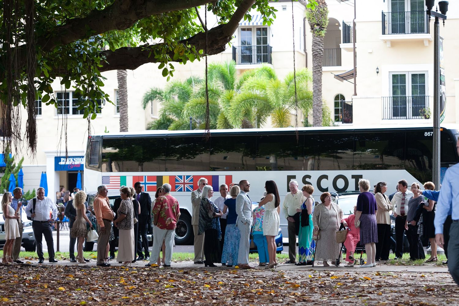 Wedding guests lining up beside white Escot charter bus outside tropical venue with palm trees.