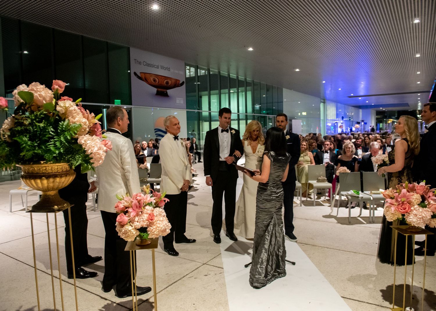 Elegant gala event with guests in formal attire mingling among floral arrangements in a modern venue.