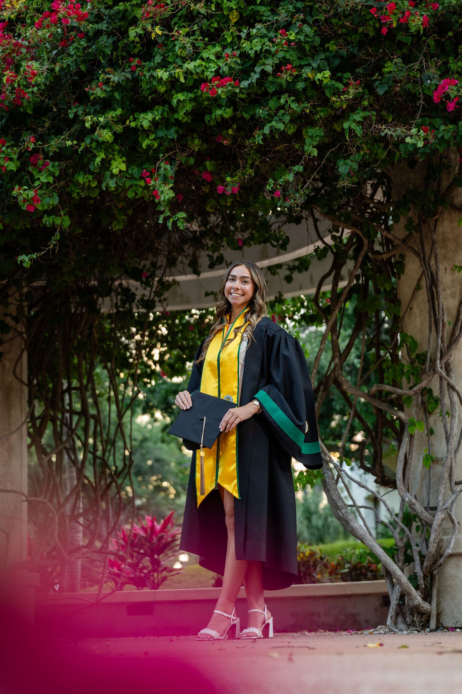 Young woman in black and gold graduation gown holding cap, smiling outdoors under flowering vine archway.