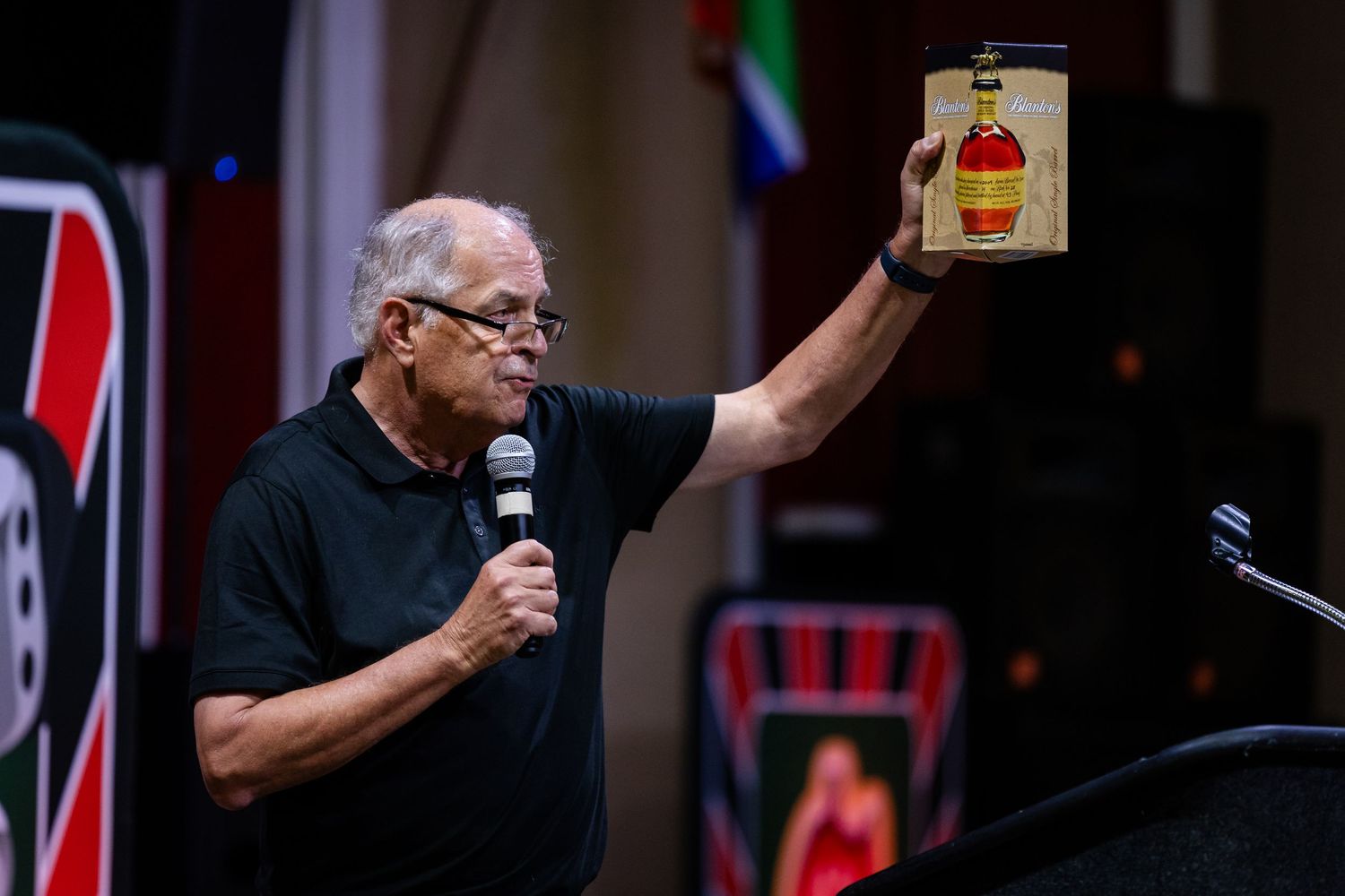 Man holds up bottle of liquor while speaking into microphone at event.
