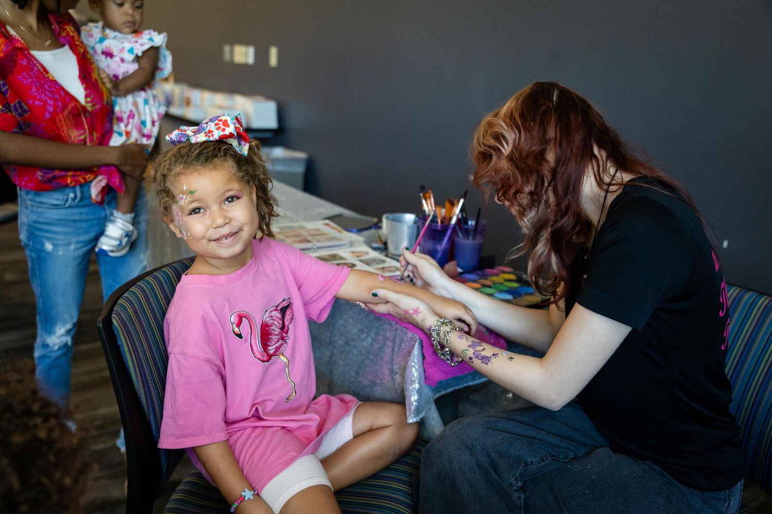 A young girl in a pink shirt gets her hand painted by a face painter at a children's party event.