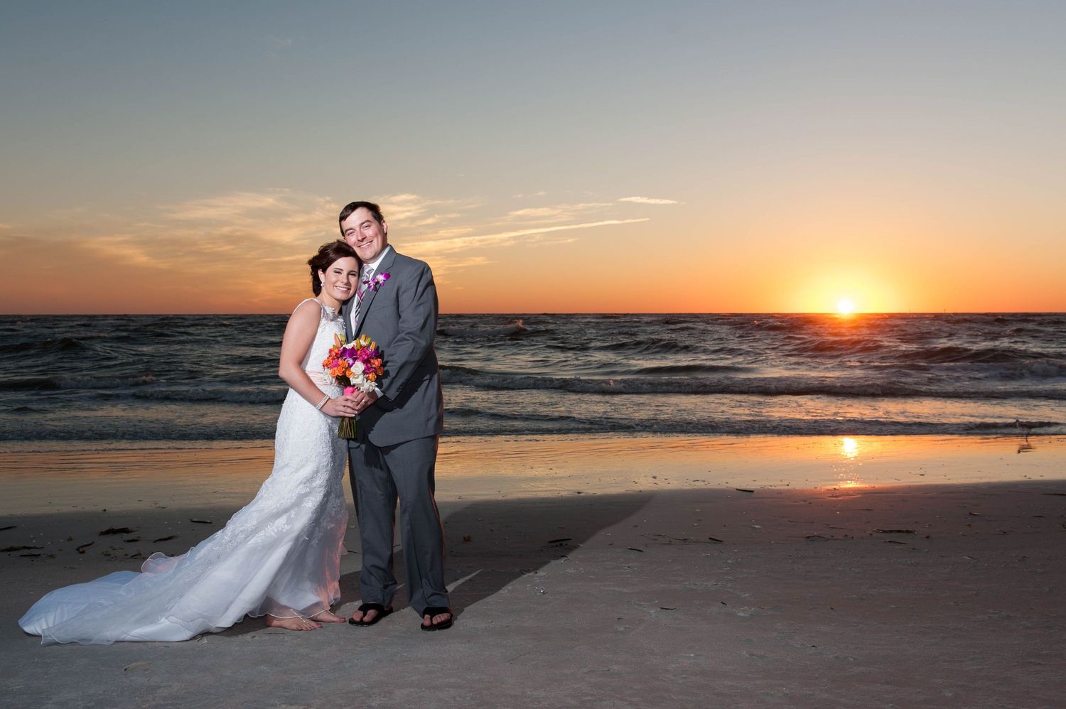 Bride and groom embrace on beach holding colorful bouquet with stunning golden sunset over ocean behind them.