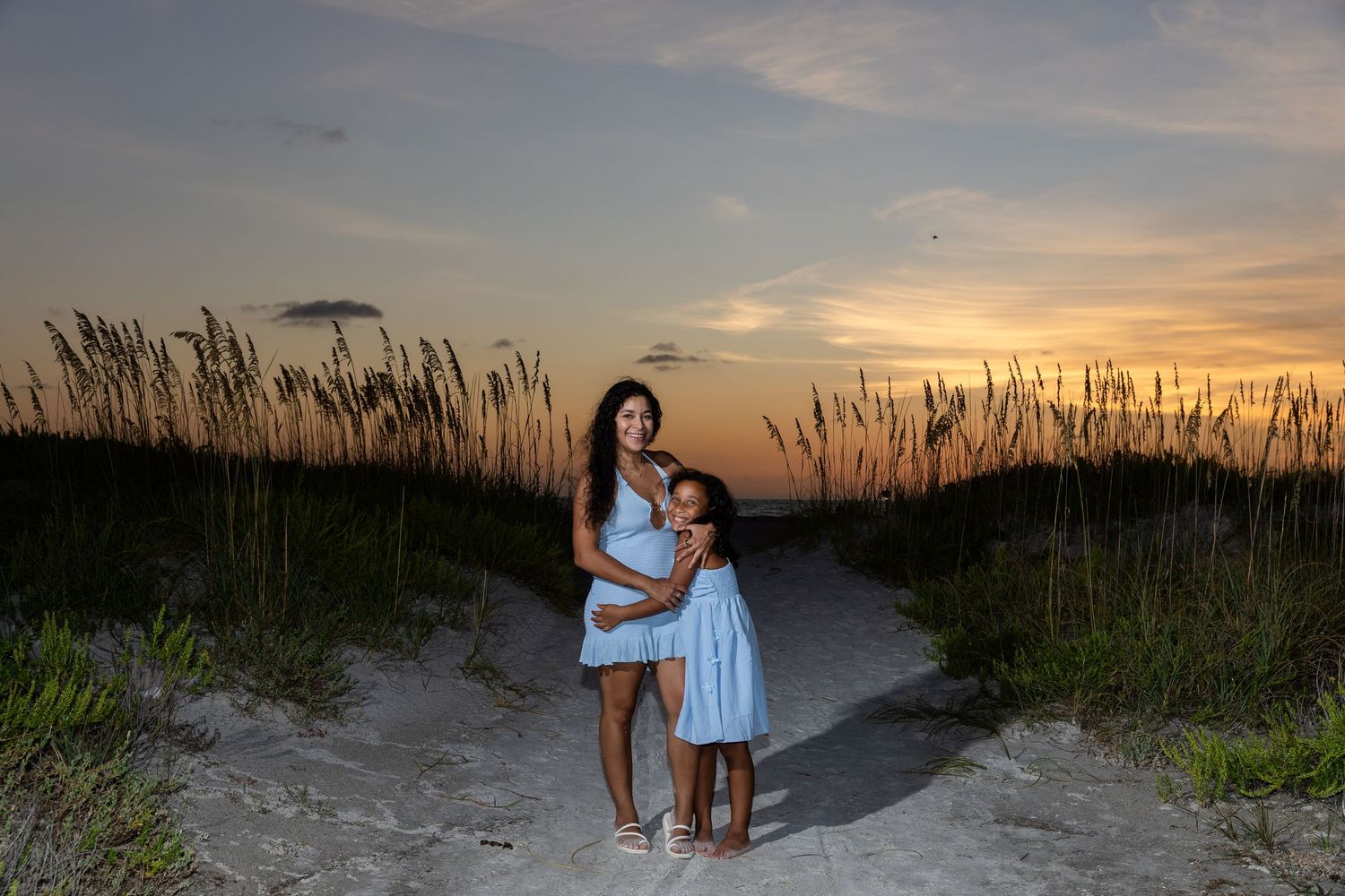 Mother and daughter in blue dresses pose on a sandy beach path at sunset with sea oats in the background.