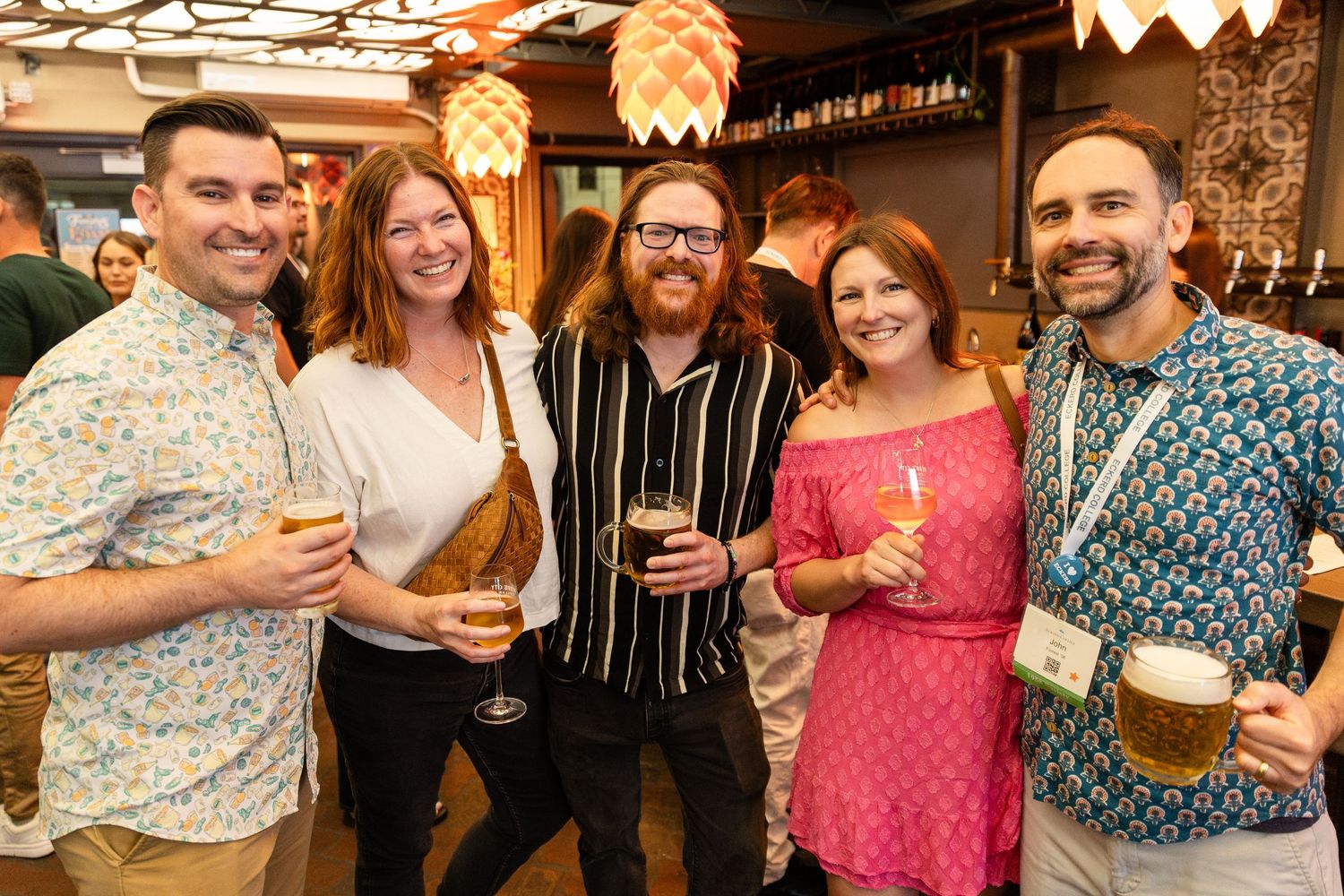Five friends smiling and holding beers at a lively bar social event.