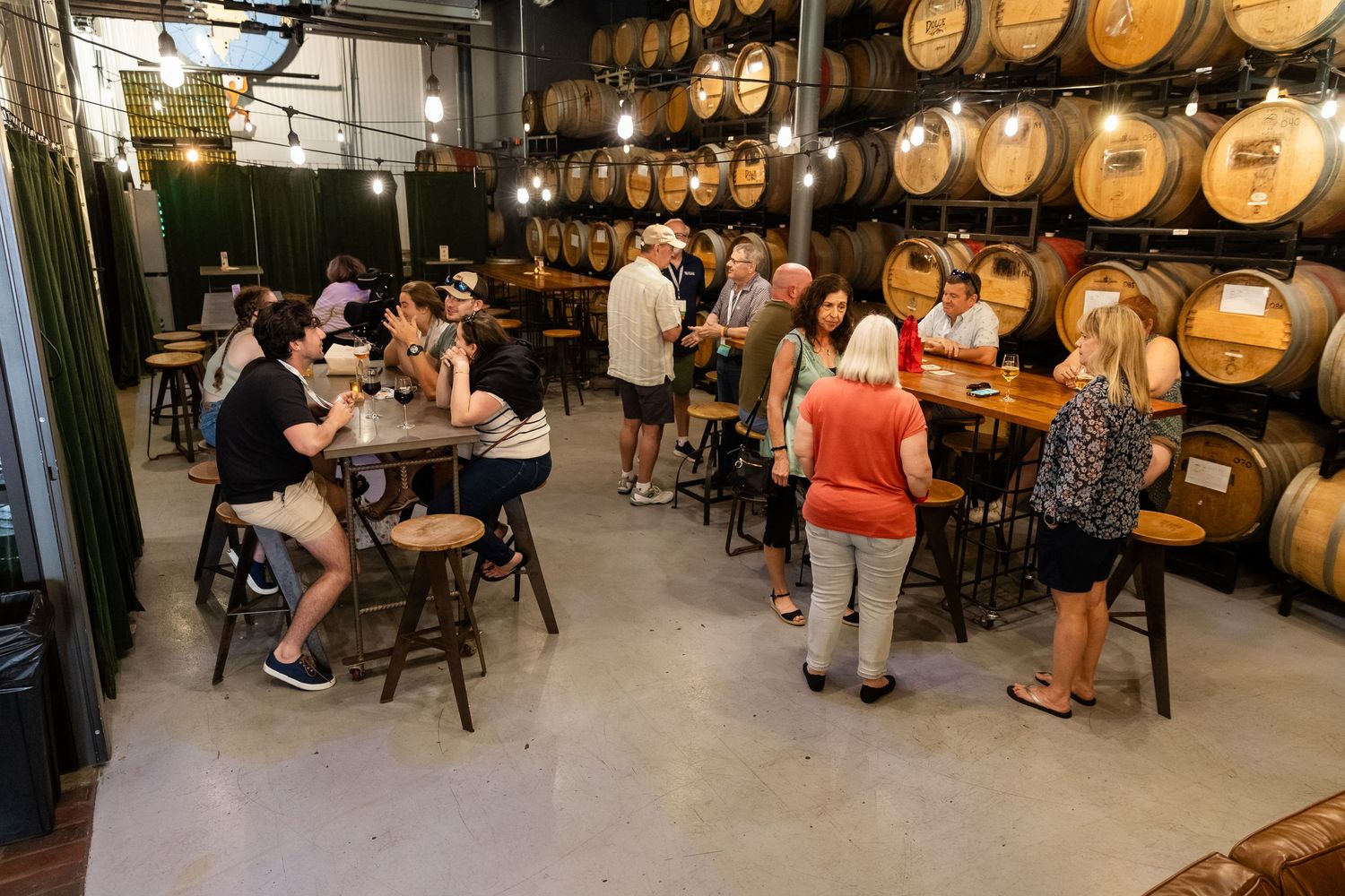 Guests socializing in a rustic brewery tasting room surrounded by wooden barrel walls.