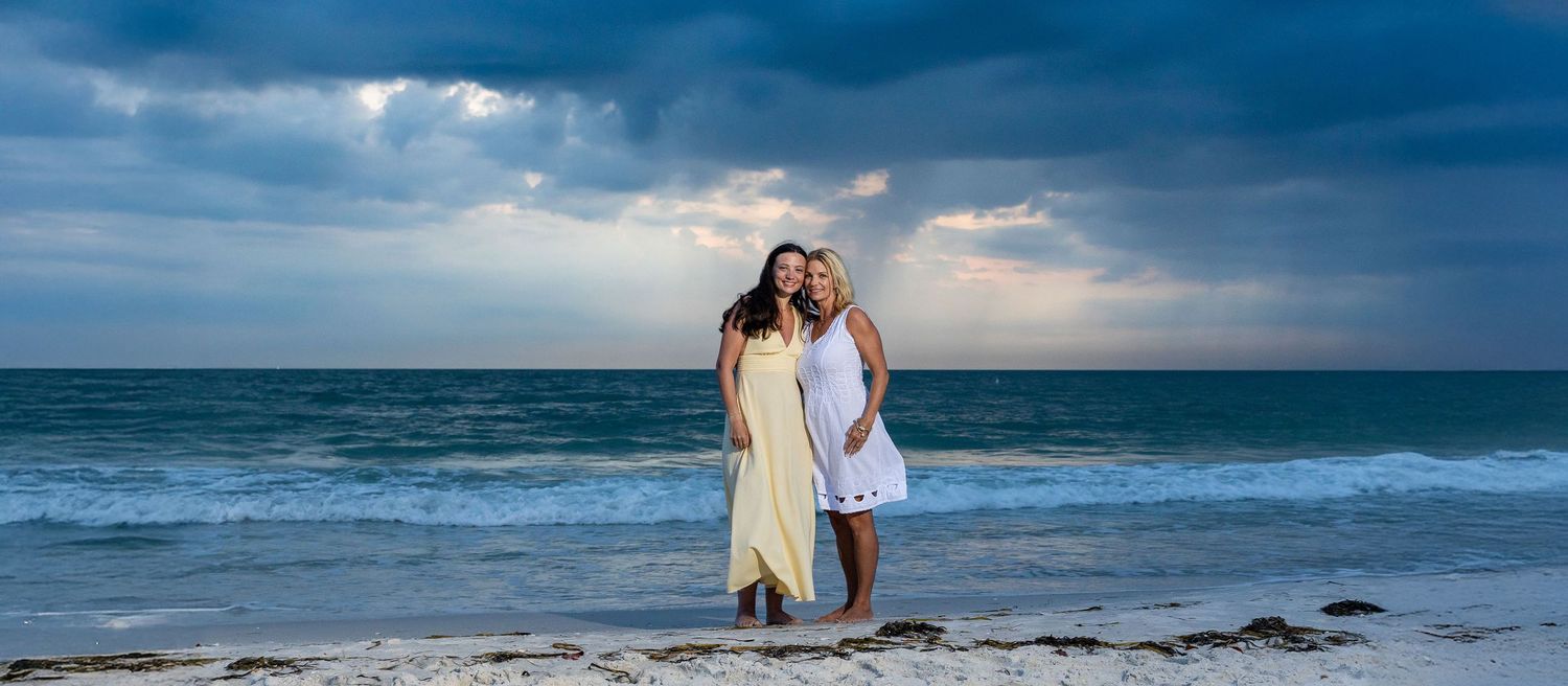 Two women posing on a sandy beach with dramatic cloudy skies and ocean waves in the background.