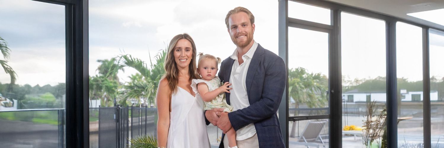 A happy young family with a baby poses indoors near large windows with tropical greenery visible outside.