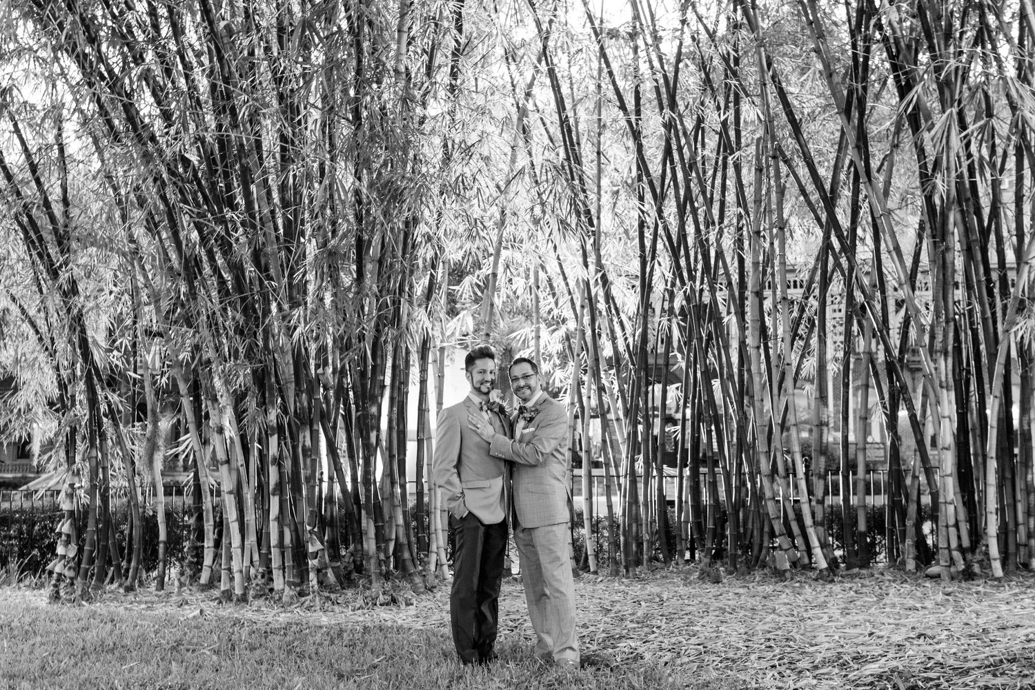 Two men embrace affectionately in a black and white photo surrounded by tall bamboo trees outdoors.