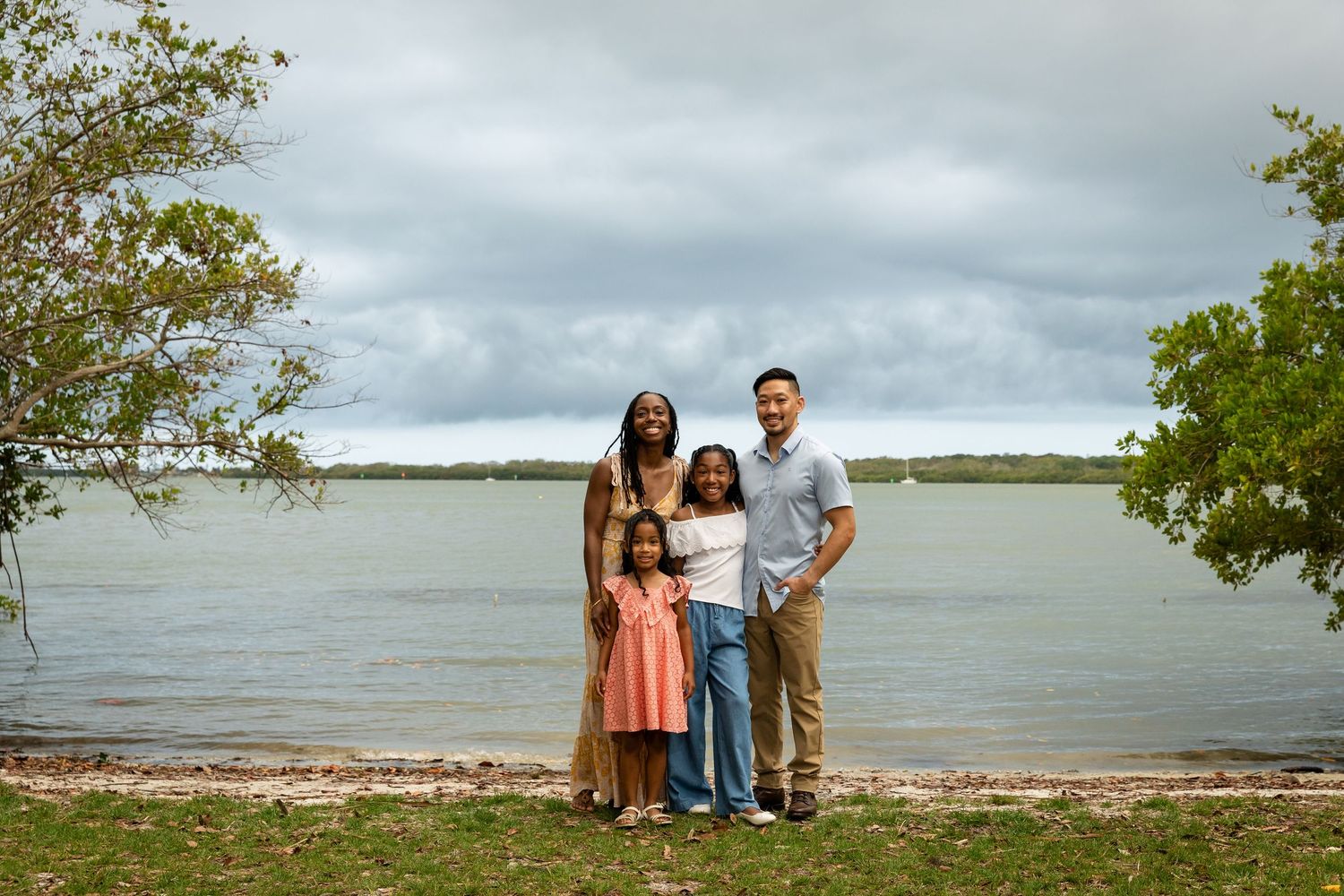 Family of four posing together on a scenic waterfront beach with lush trees and cloudy skies.