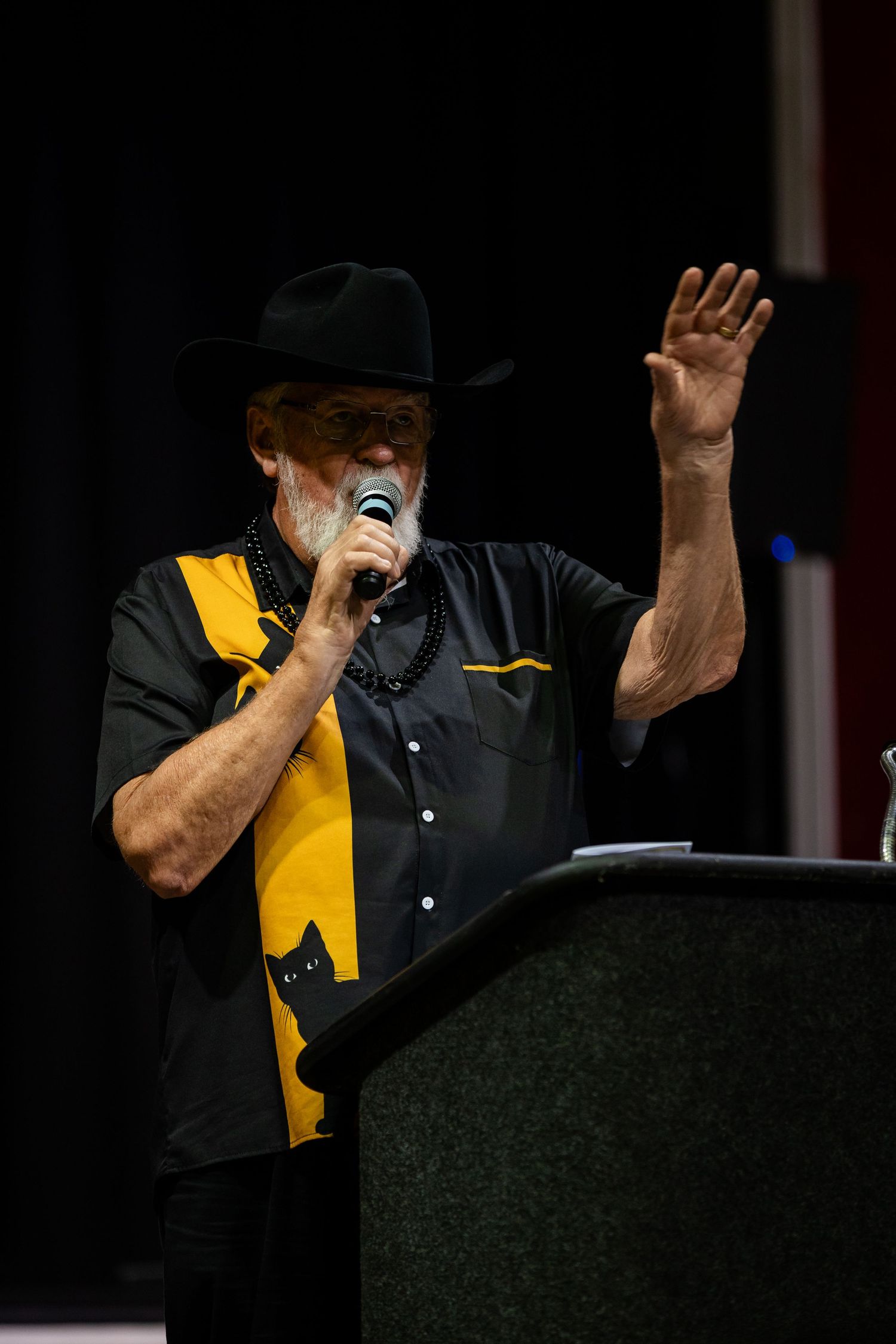 Man in black cowboy hat and yellow shirt speaks into microphone while raising hand at podium.