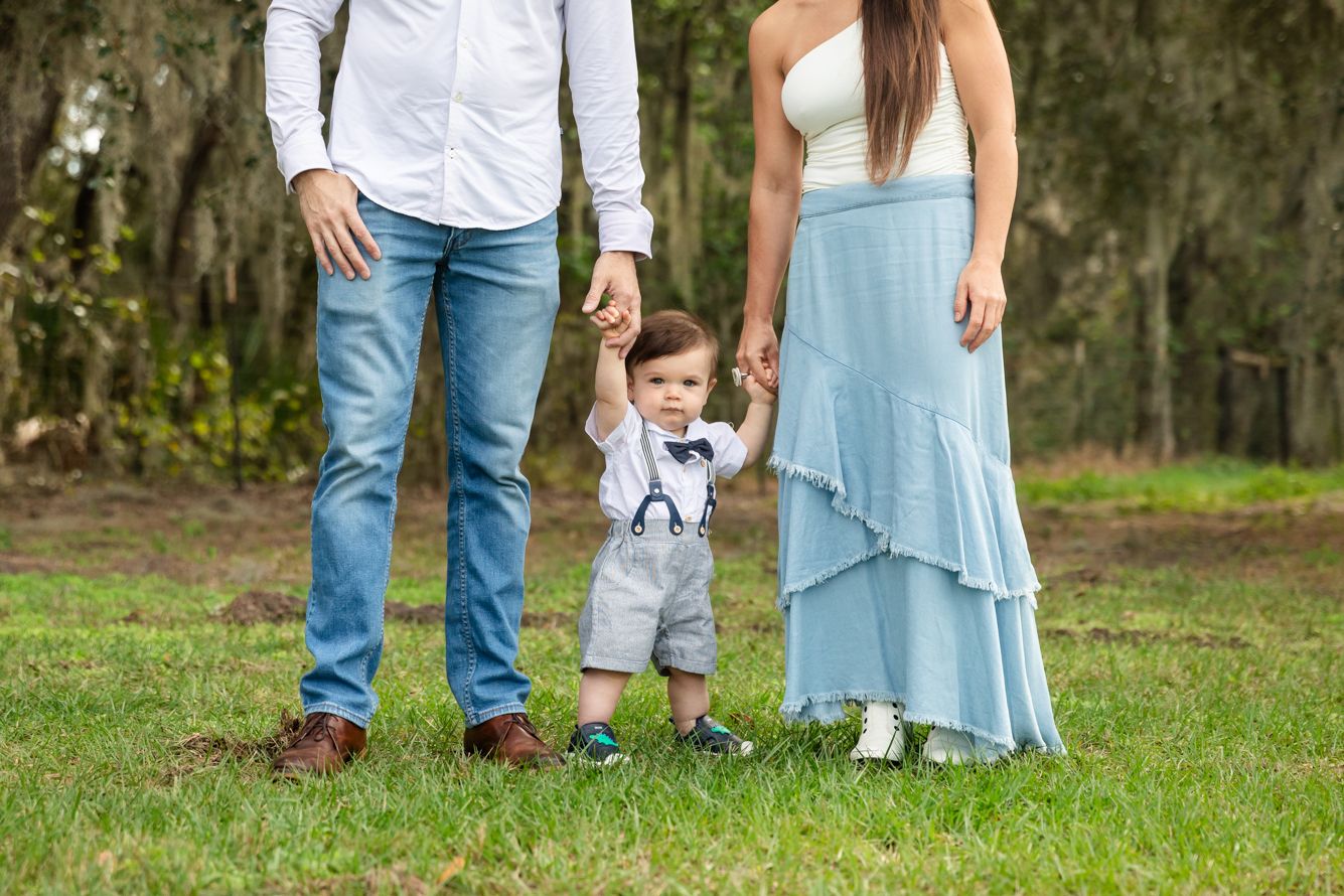 Family photo of parents holding hands with toddler boy in suspenders on green grass outdoors.