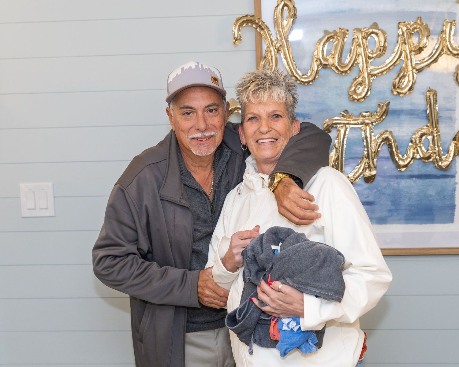 Smiling couple posing together indoors in front of a Happy Birthday balloon sign, woman holding a baby.