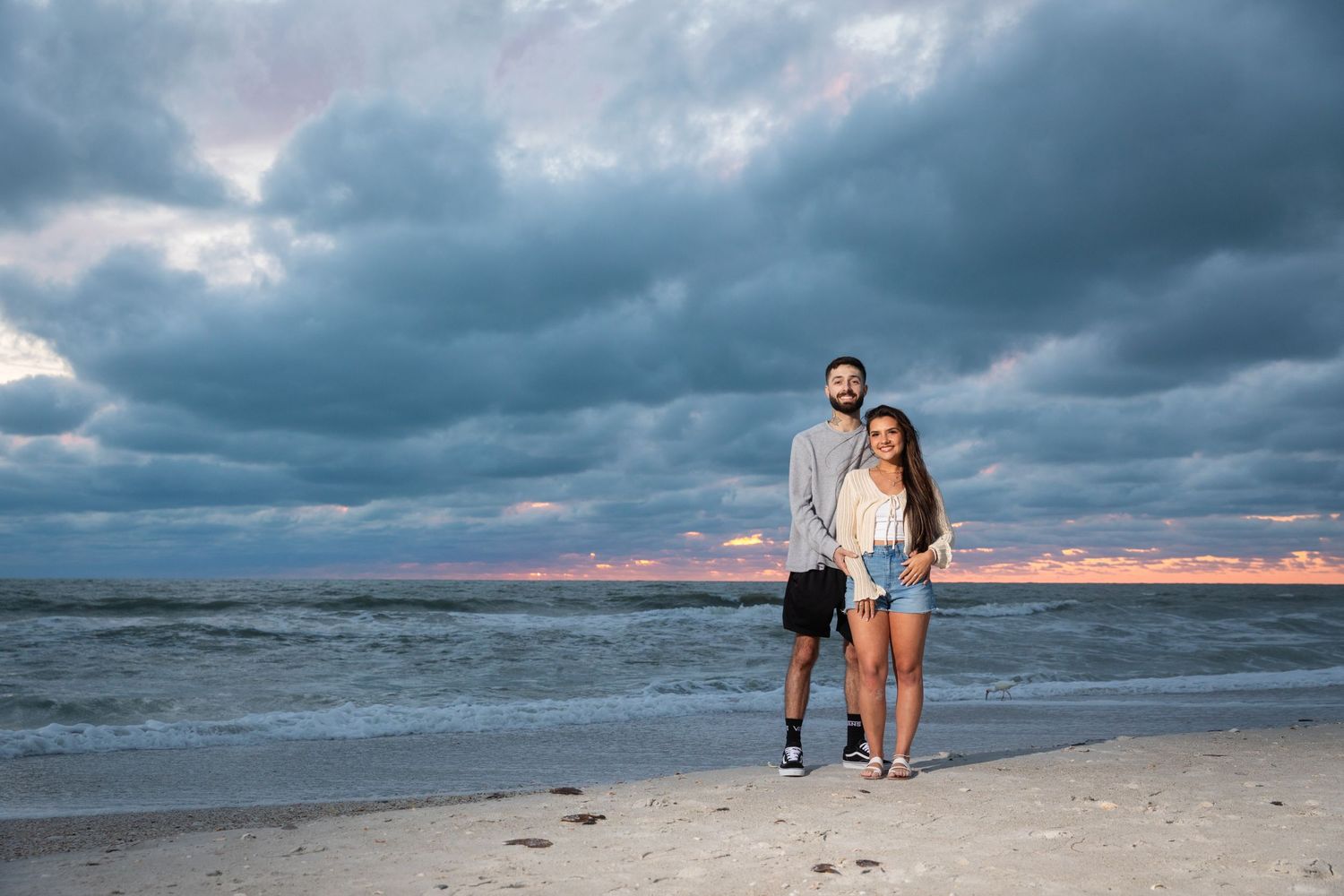 Couple posing together on a sandy beach at sunset with dramatic cloudy sky over the ocean.