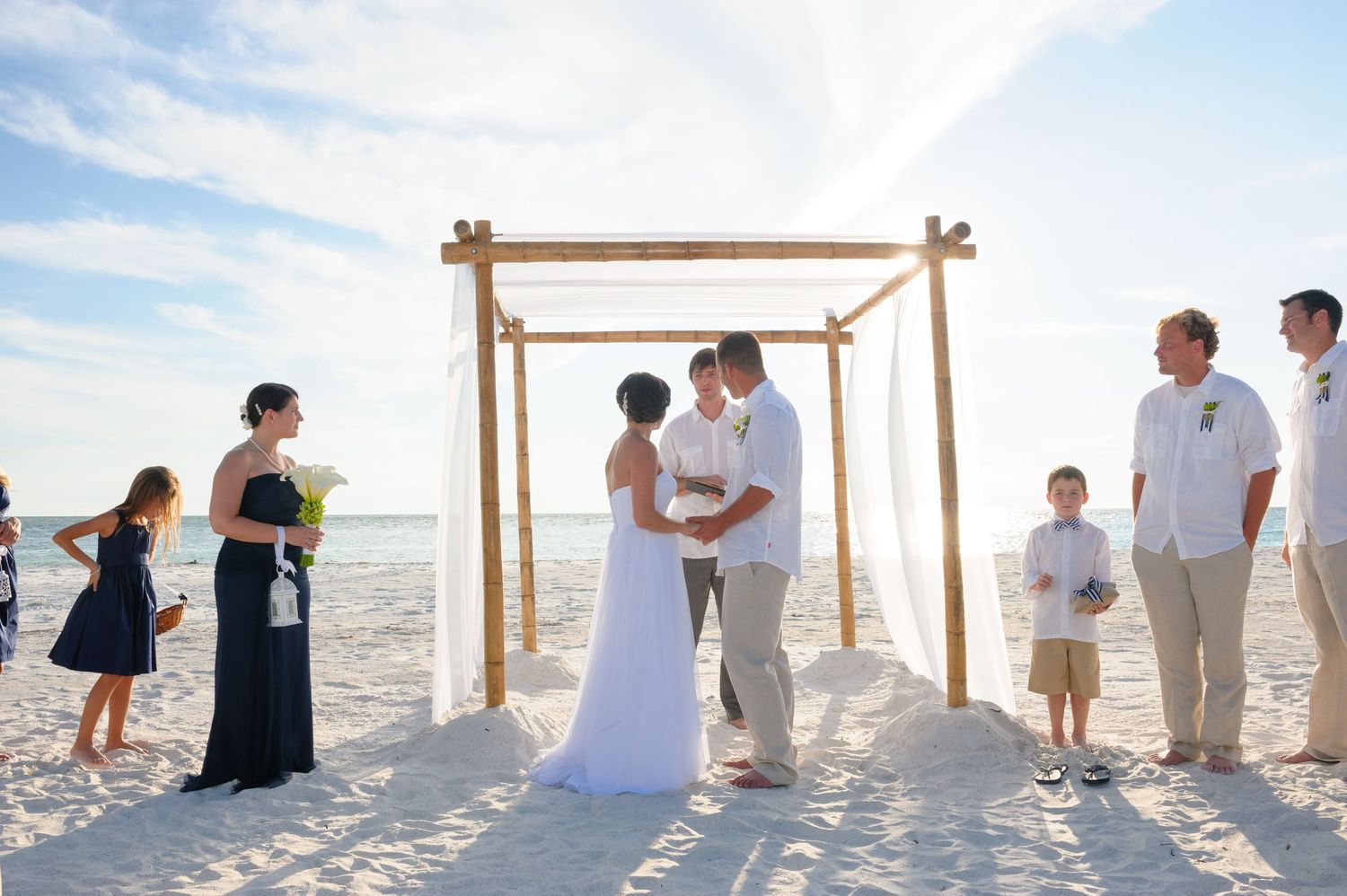 Couple kissing at a romantic beach wedding ceremony under a wooden arch with wedding party and guests on white sand.