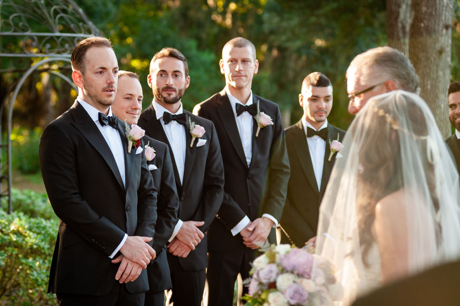 Groomsmen in black tuxedos with bow ties stand at outdoor wedding ceremony, watching bride approach the altar.