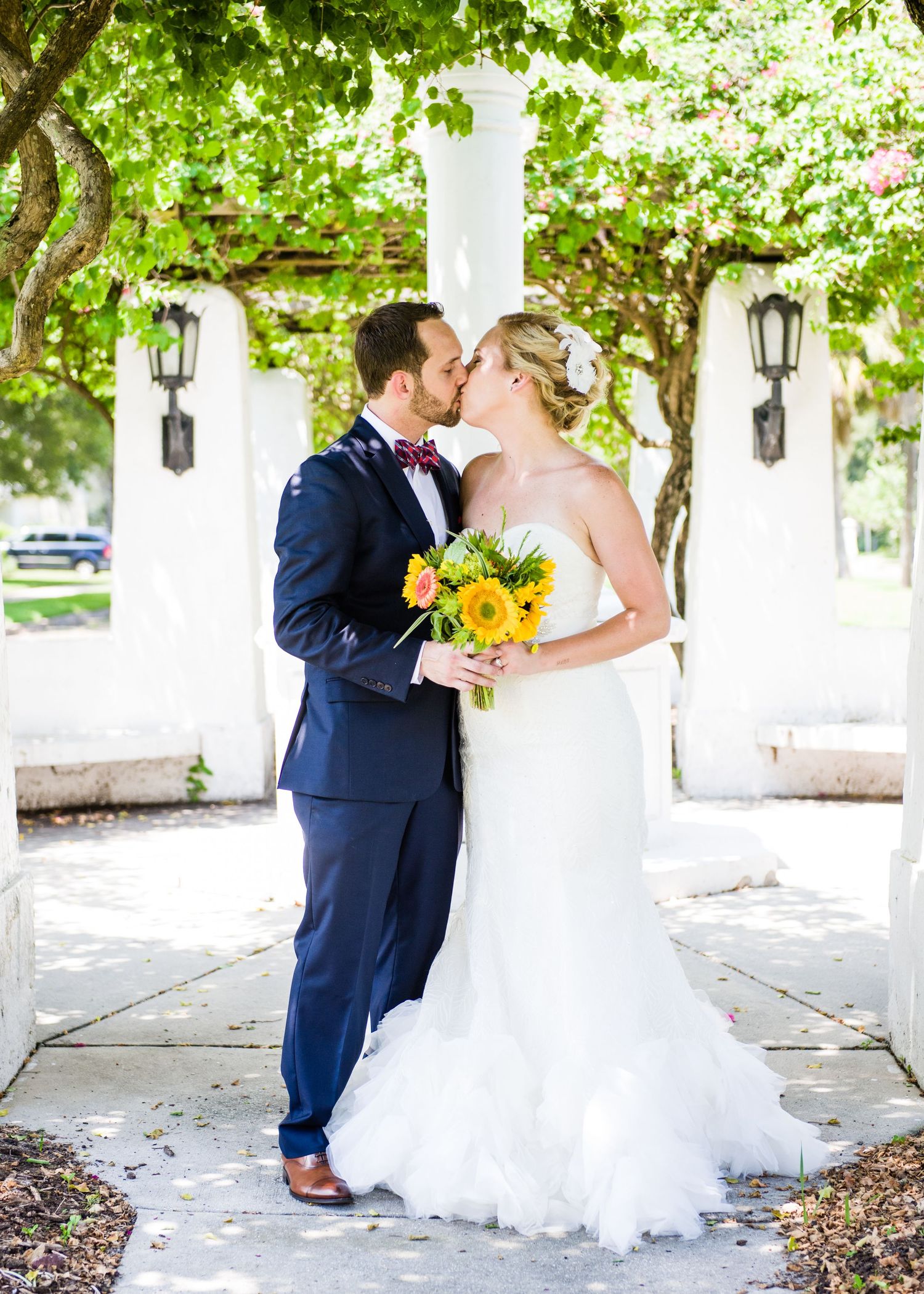 A bride and groom share a kiss outdoors, she holds a sunflower bouquet, he wears a navy suit with bow tie.