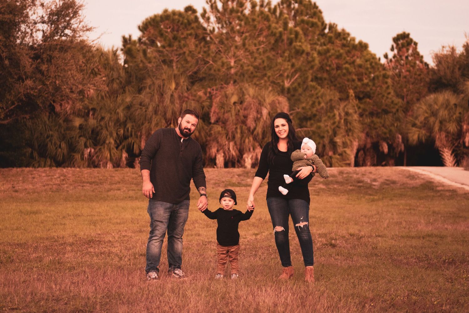 Family of four posing outdoors in a grassy field with trees, parents holding toddler's hands while mom holds baby.