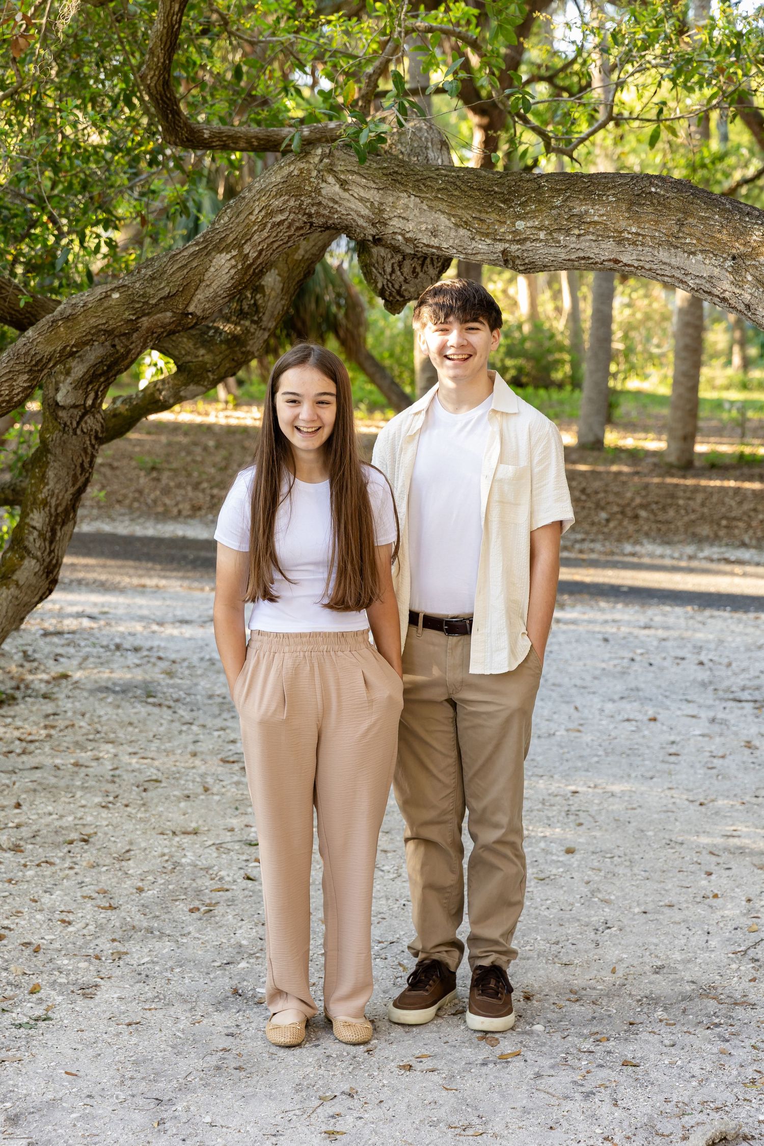 Two smiling teenagers in neutral tones stand together under a large oak tree in a sunny outdoor park setting.