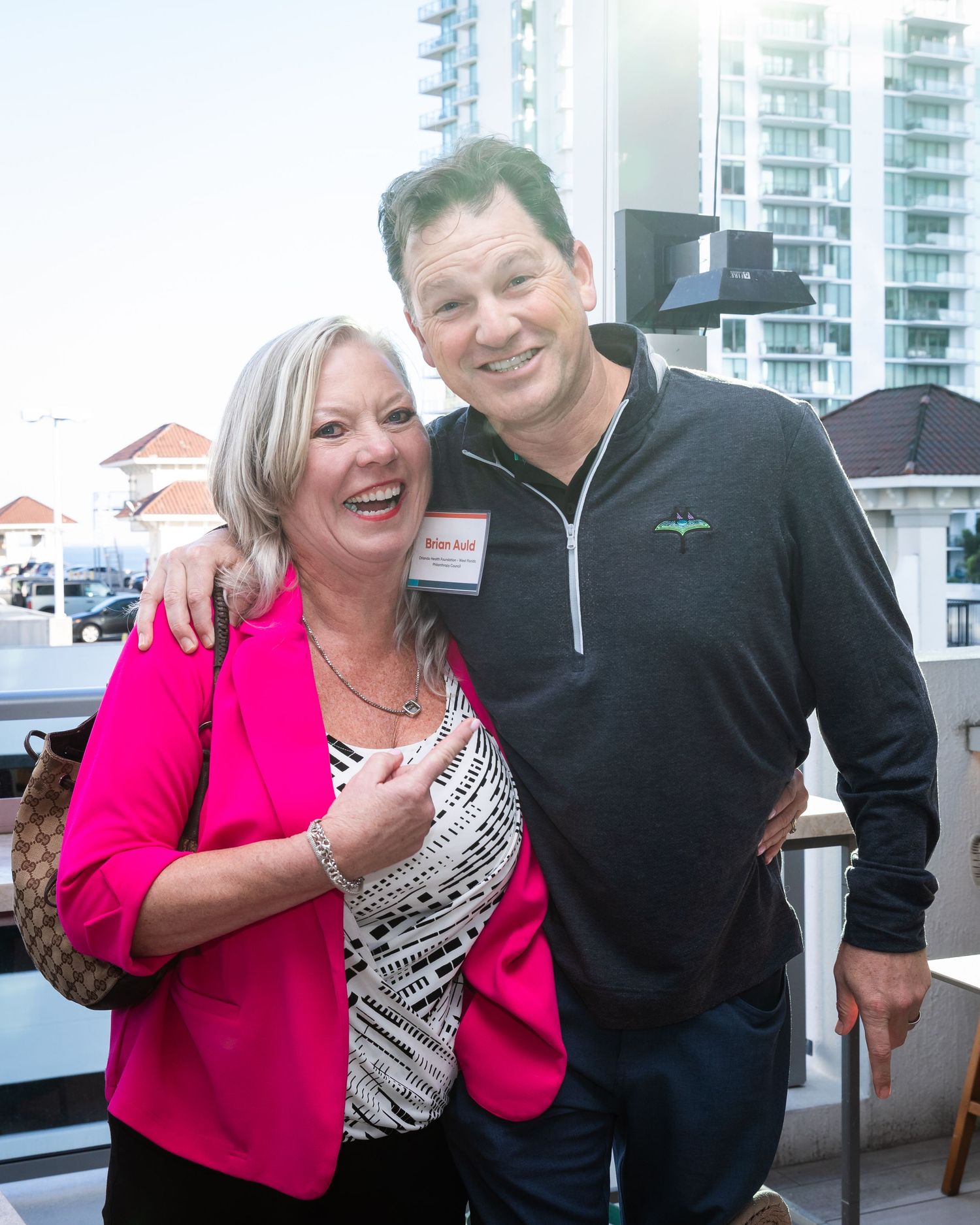 A smiling woman in a pink blazer poses with a man in a black zip-up jacket at a networking event.