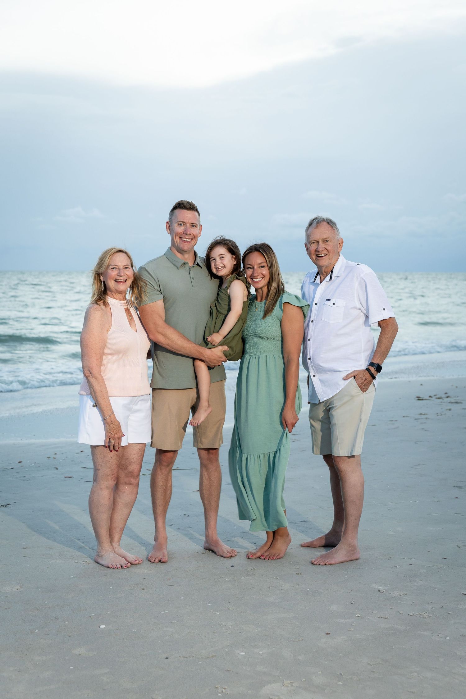 Multi-generational family of five posing together on a sandy beach with ocean waves in the background.