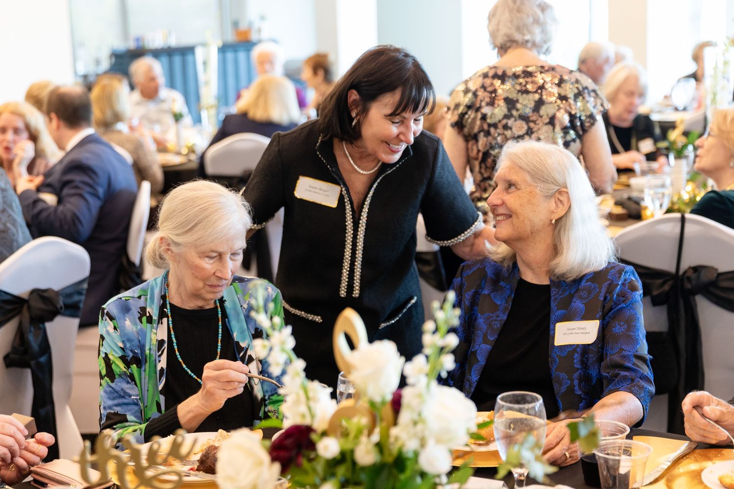 Elderly women smile at a formal banquet event while a younger woman in a name tag leans in to chat.