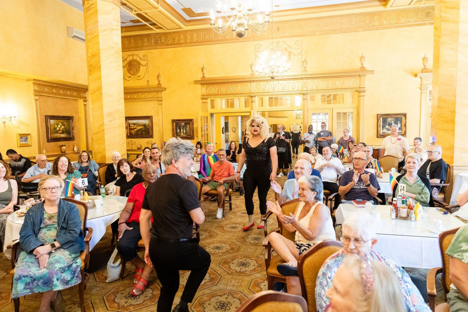 Performers entertaining a seated crowd of seniors at a vibrant indoor Pride celebration in a grand ballroom.