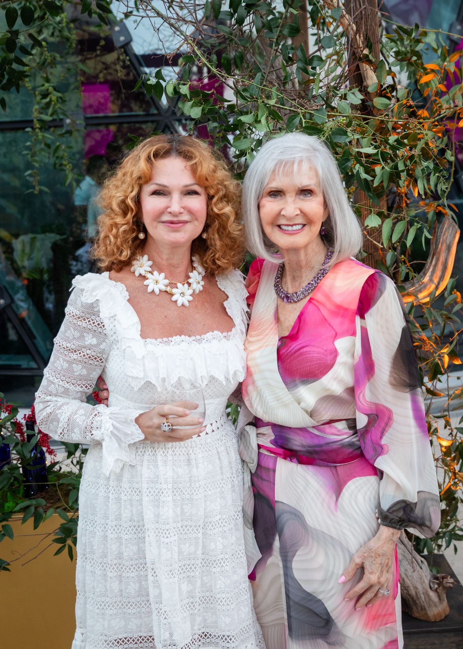 Two elegantly dressed women posing together outdoors, one in white lace dress and one in colorful pink wrap dress.