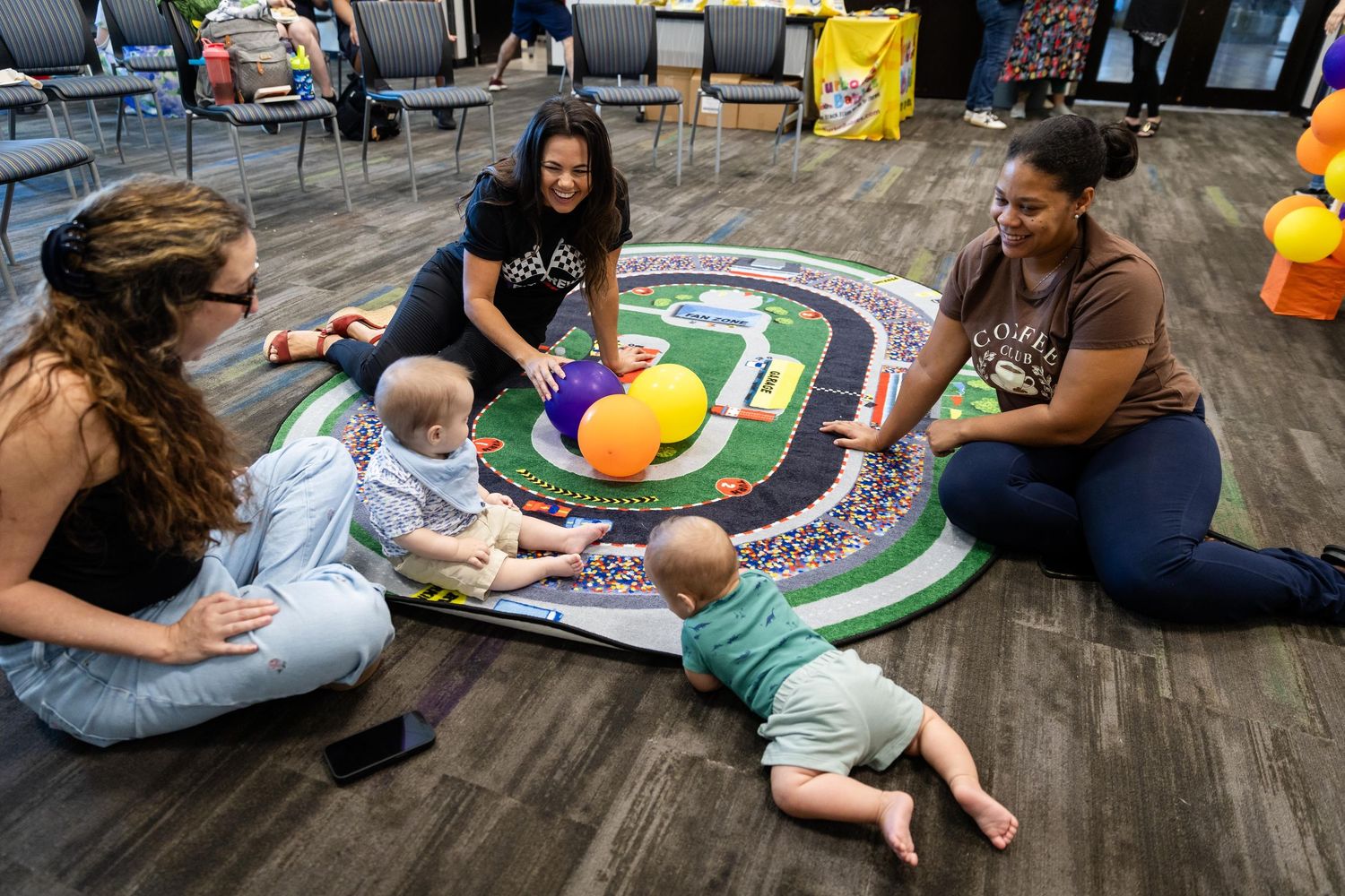 Two women and babies playing on a colorful racetrack mat with balloons in a bright indoor space.