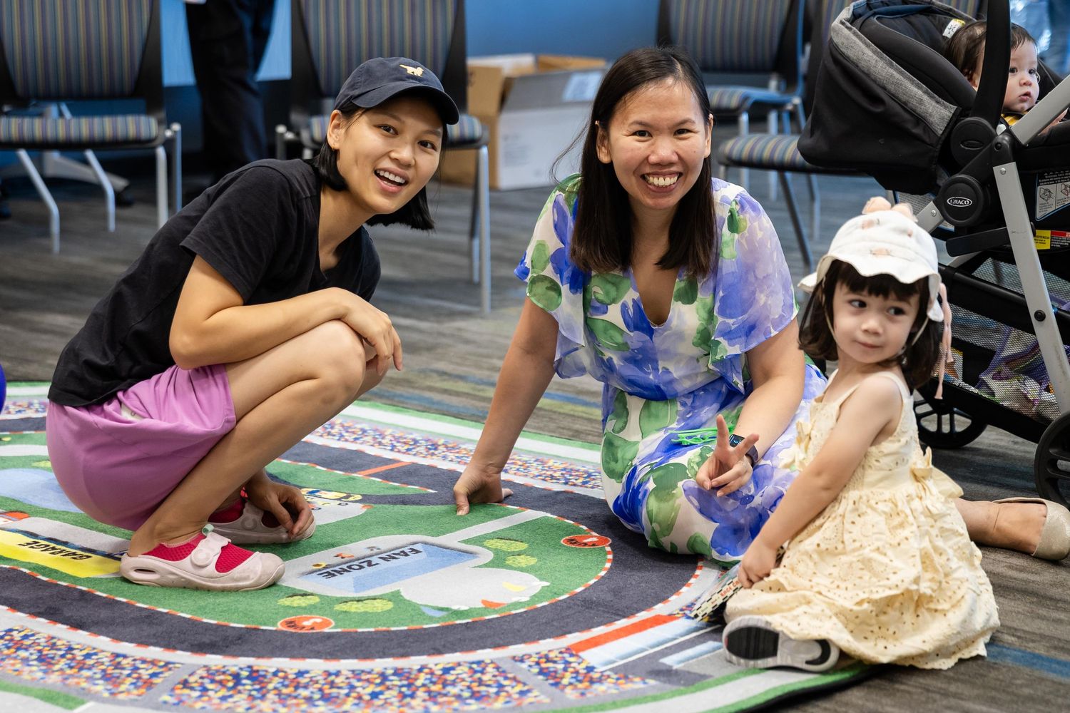 Two women and a young girl smile together on a colorful play mat in a bright indoor space.