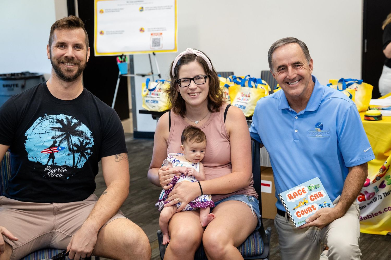 Family with baby poses alongside man holding children's book at community book donation event.