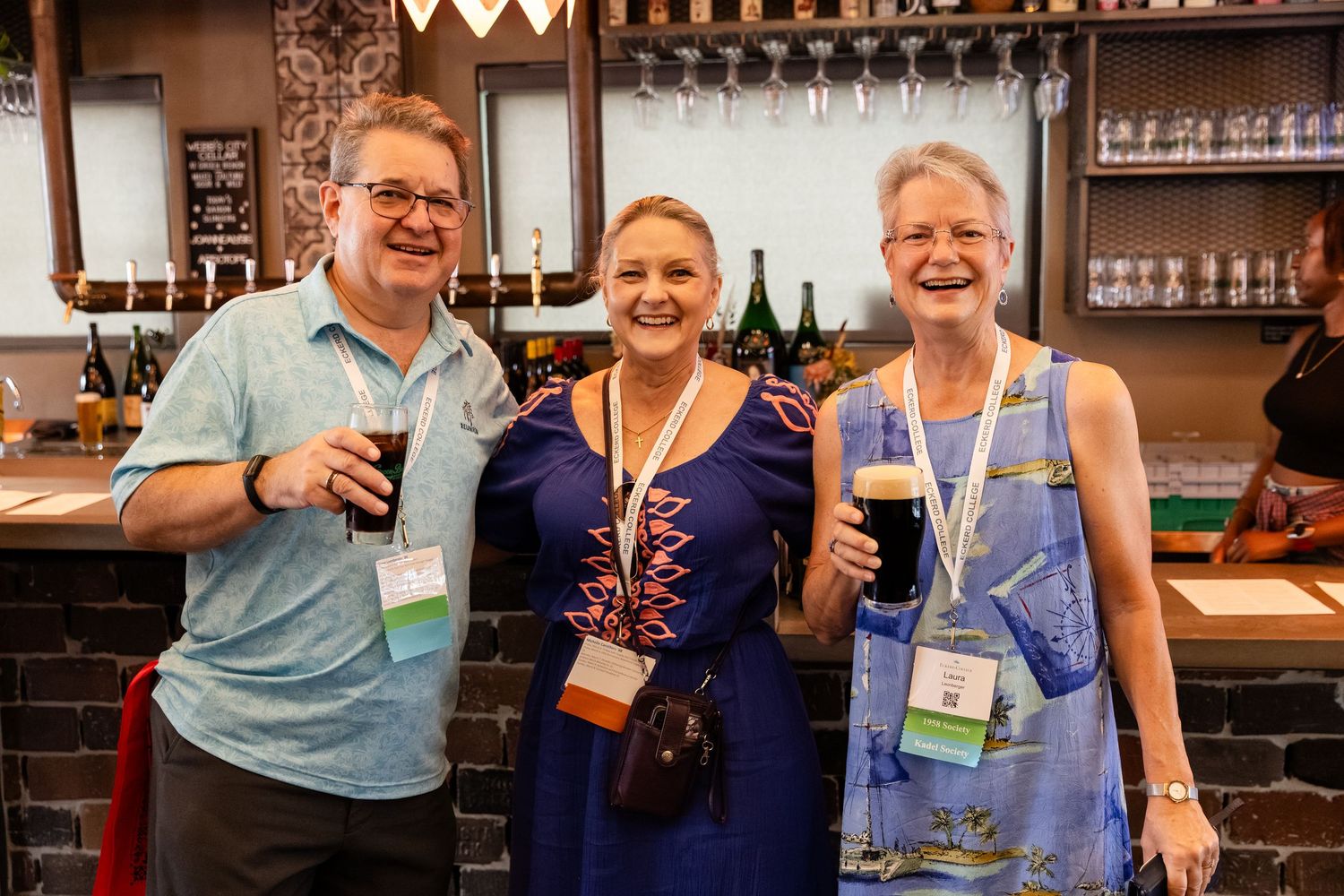 Three smiling adults holding dark beers at a bar, wearing lanyards at what appears to be a social event.