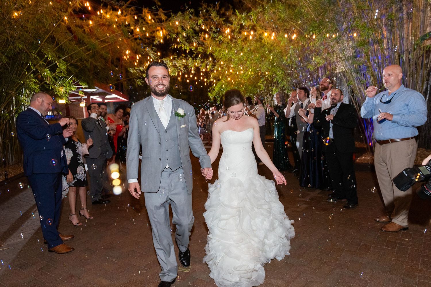 Newlywed couple walks hand in hand through cheering guests under twinkling lights at outdoor wedding reception.