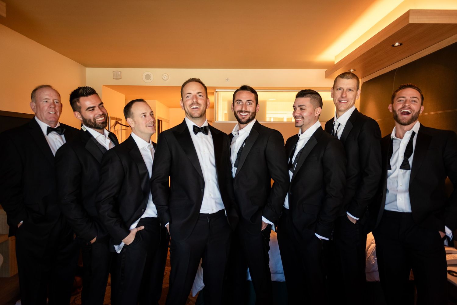 Groom poses with seven groomsmen all wearing classic black tuxedos indoors before wedding ceremony.