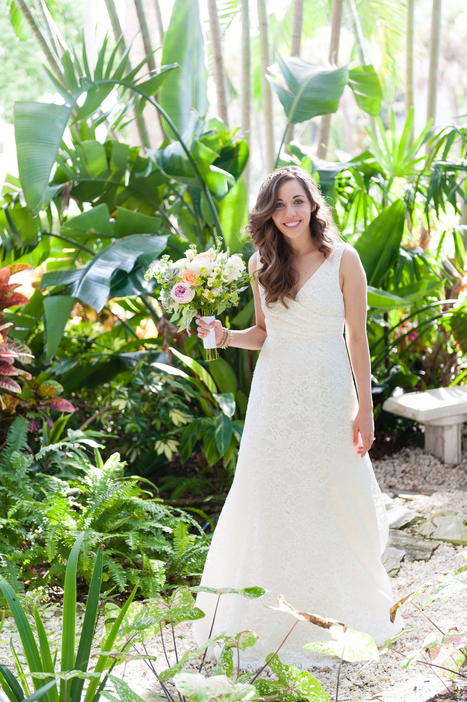 Smiling bride in white lace A-line gown holding colorful bouquet in lush tropical garden setting.