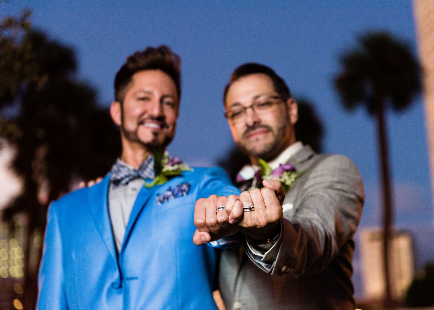 Two grooms in suits show off wedding rings, smiling outdoors at dusk with palm trees in background.
