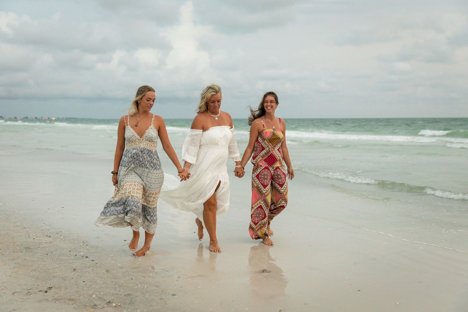 Three women in colorful summer dresses walking barefoot along a white sandy beach with ocean waves.