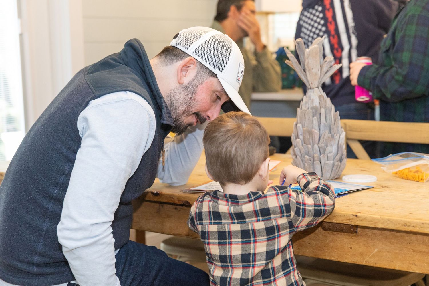 Man in trucker hat bends down to interact with young boy at wooden table with decorative fish.