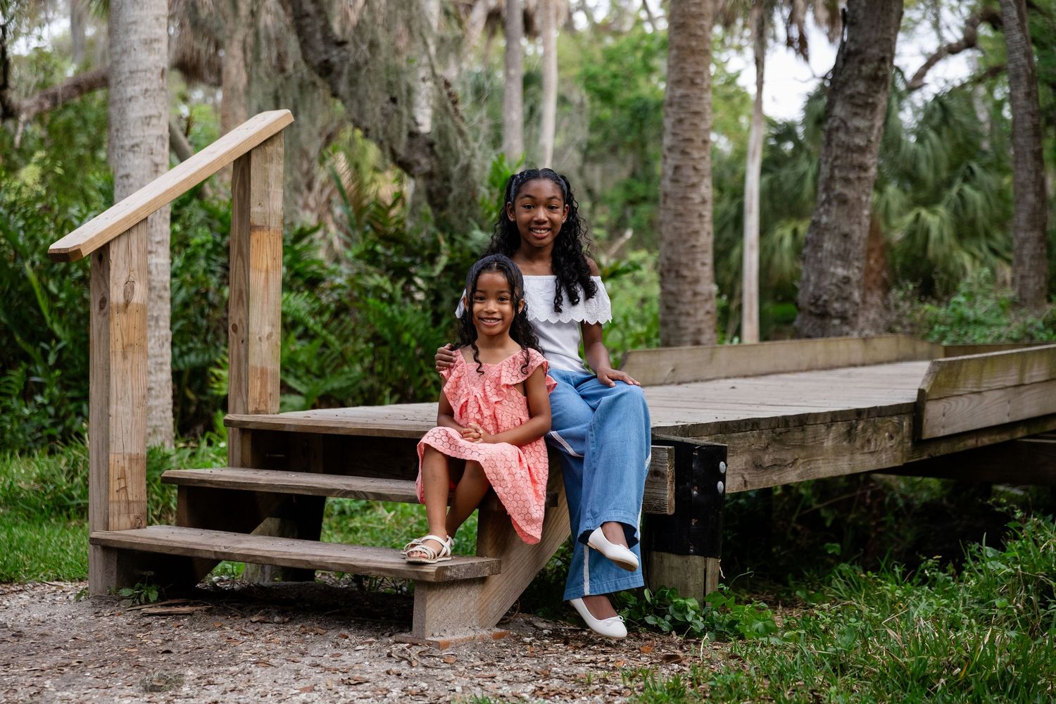 Two young sisters sitting on wooden park steps surrounded by palm trees and lush greenery.