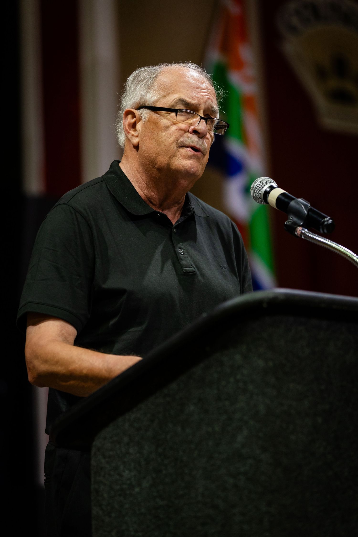 Elderly man in black polo shirt speaking at a dark podium with microphone at a formal event.