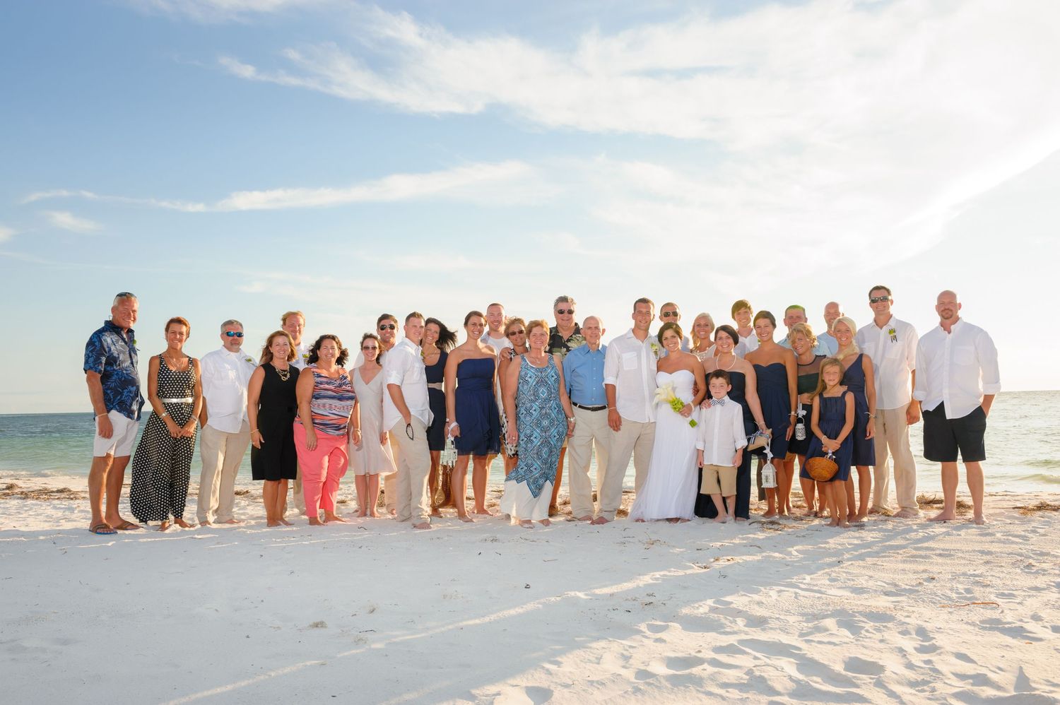 Large wedding party posing together on a white sandy beach with clear blue skies and calm ocean waters in the background.