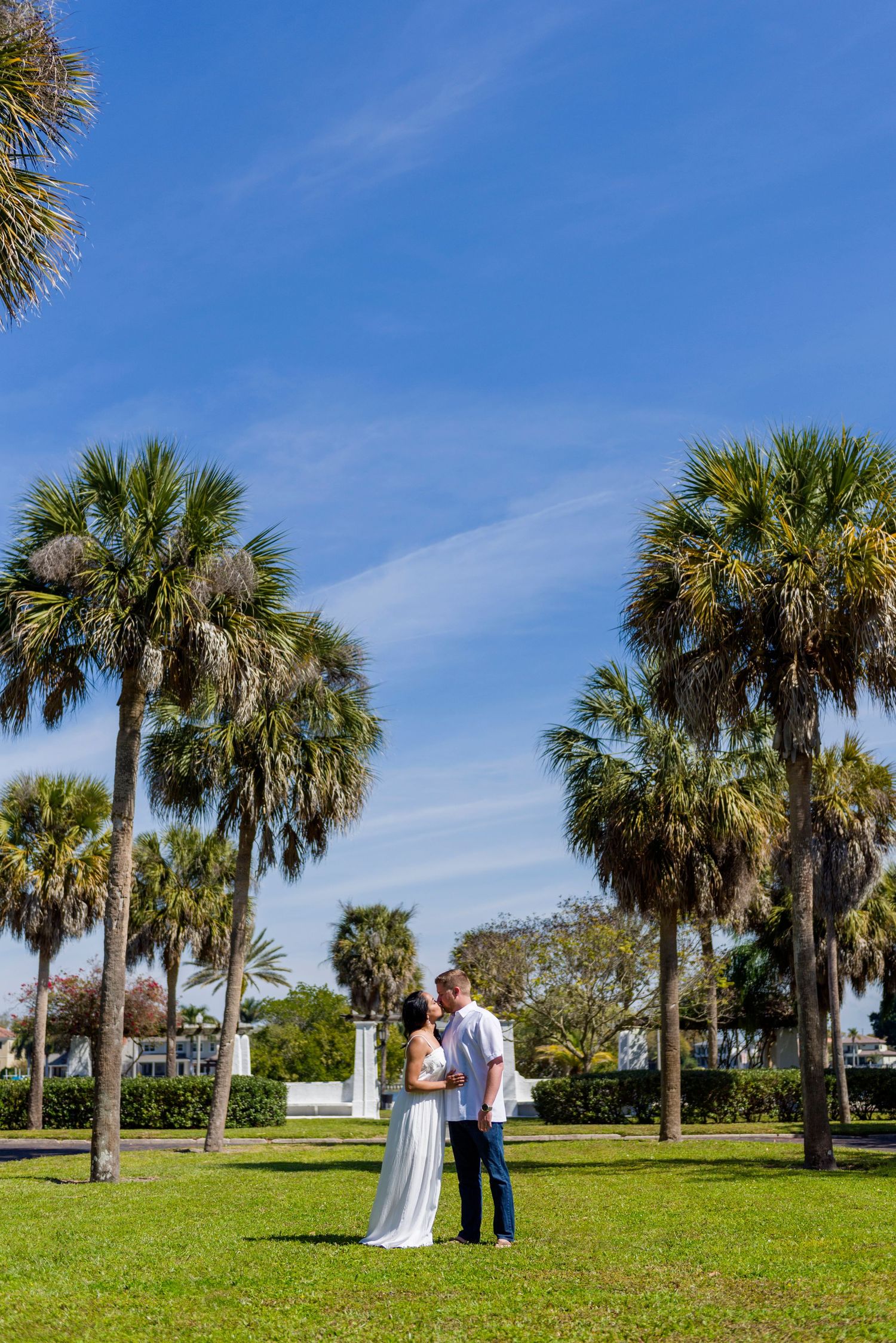 Couple embracing on green grass surrounded by tall palm trees under a bright blue sky.