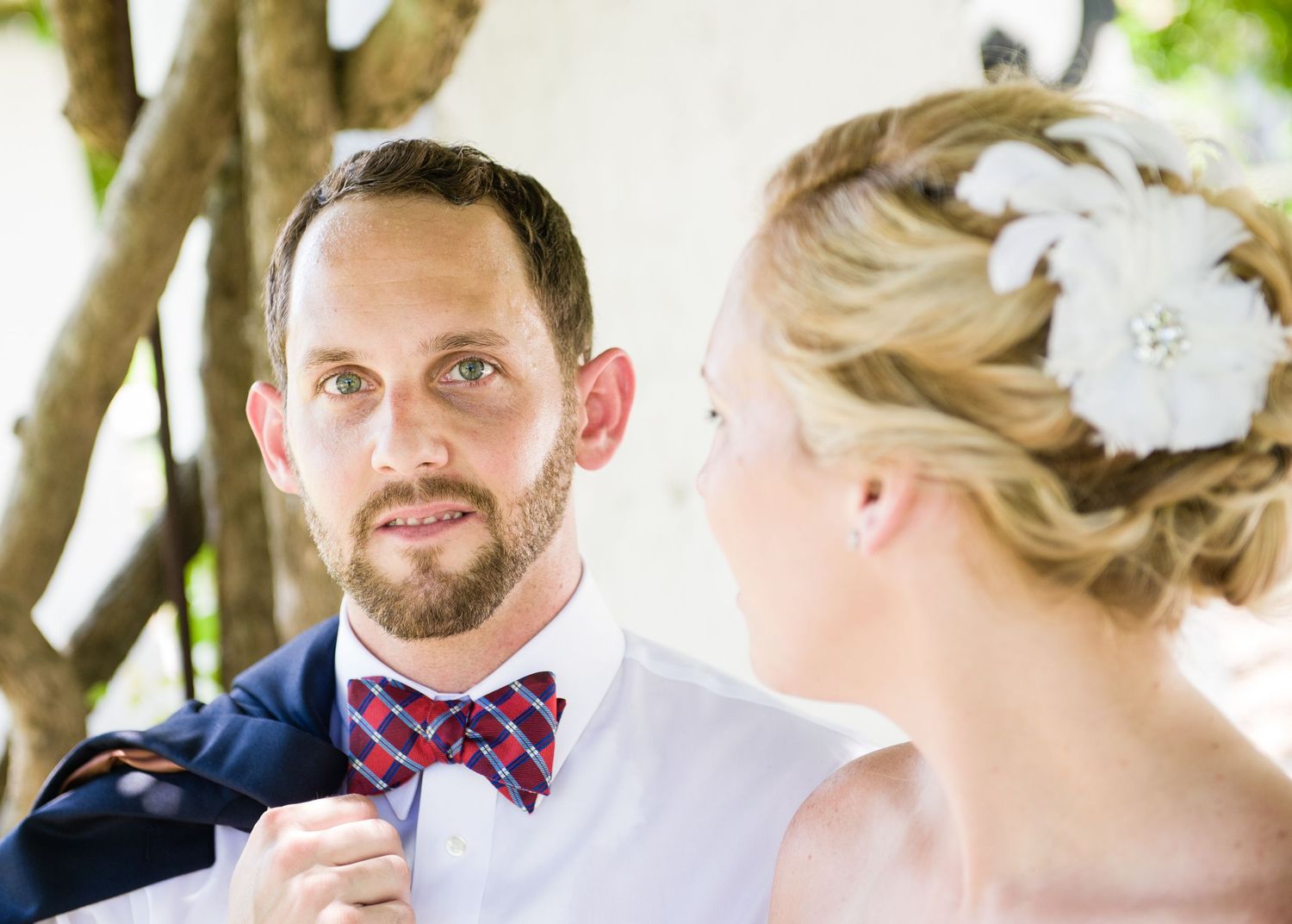 Groom in blue suit and red plaid bow tie gazes at blonde bride wearing white floral hair accessory outdoors.